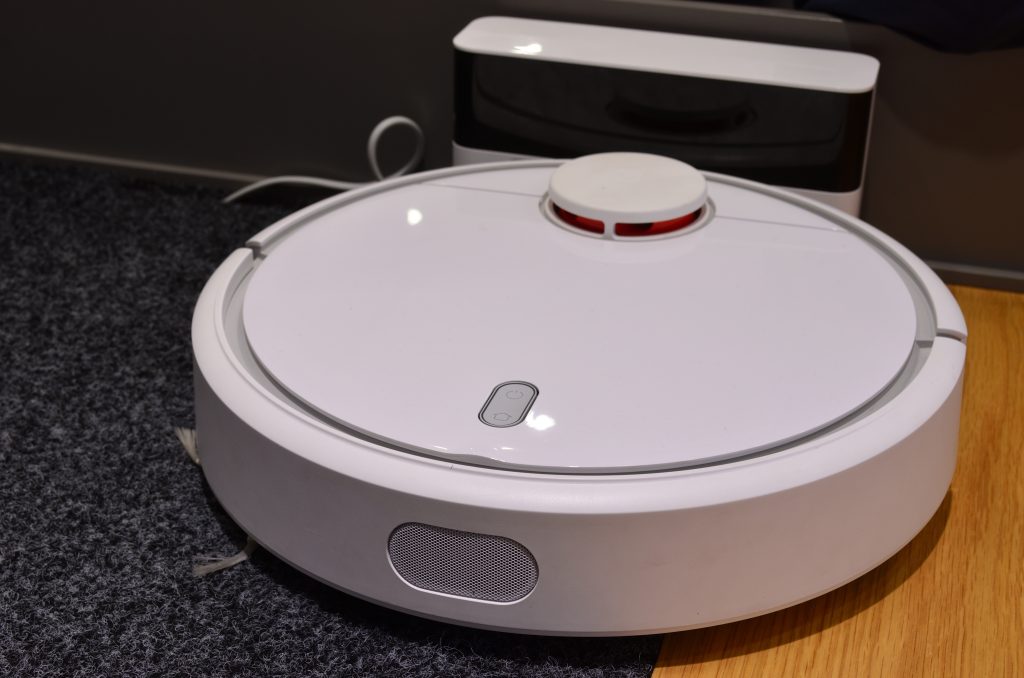 Robot aspirapolvere bianco sul passaggio tra tappeto e parquet, con la base di ricarica sullo sfondo